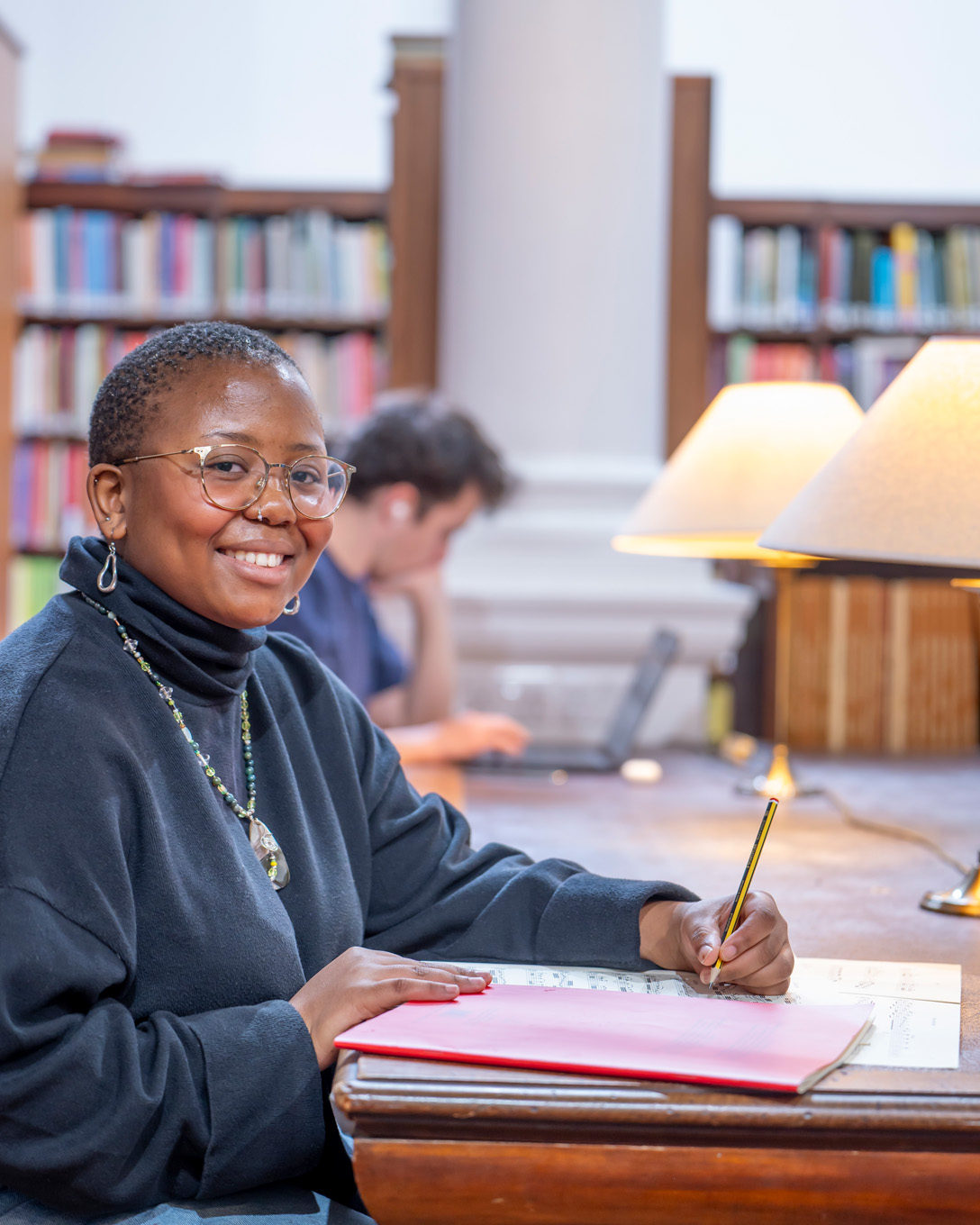 A black women, wearing a dark jumper, writing and sitting in a library desk.